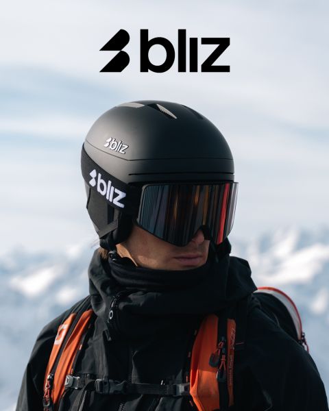 Une personne portant un casque de ski noir et des lunettes de ski à effet miroir, tous deux portant l'inscription "Bliz", se tient à l'extérieur avec des montagnes enneigées en arrière-plan. Le logo Bliz est bien visible en haut de l'image.
