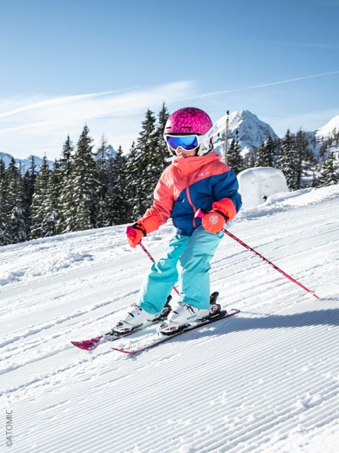 Un jeune enfant portant un équipement de ski coloré et un casque rose descend une pente enneigée à ski, avec des arbres et des montagnes en arrière-plan.