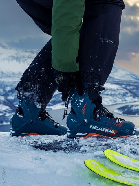 Une personne portant des chaussures de ski SCARPA ajuste les clips de ses chaussures sur un sol enneigé, avec des montagnes et des skis en arrière-plan.