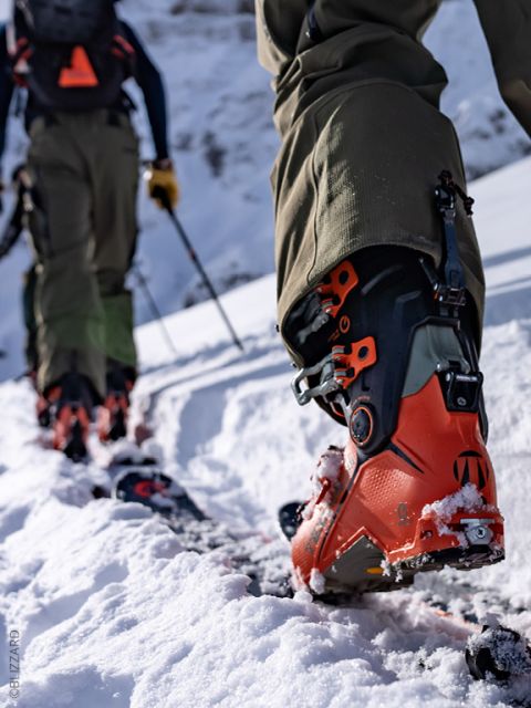 Gros plan sur deux personnes marchant dans la neige en montée avec des chaussures de ski orange et des bâtons, avec des montagnes enneigées en arrière-plan.