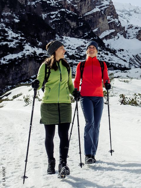 Deux femmes en vêtements d'hiver marchent avec des bâtons sur un chemin enneigé et se sourient devant un paysage de montagnes enneigées.