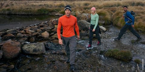 Trois personnes en équipement de plein air traversent un ruisseau rocailleux dans une région montagneuse et herbeuse, une personne devant et les autres derrière.