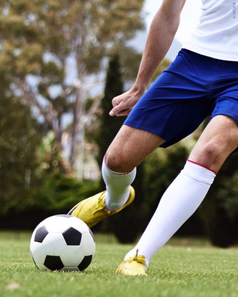 Un joueur de football en short bleu et chaussures jaunes donnant un coup de pied dans un ballon de football noir et blanc sur une pelouse, avec des arbres en arrière-plan.