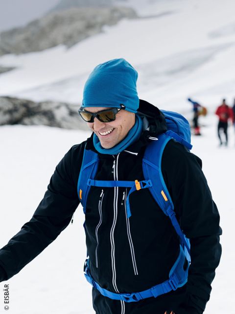 Une personne souriante en vêtements d'hiver se promène dans un paysage enneigé, portant des lunettes de soleil, un bonnet bleu et un sac à dos bleu, tandis que d'autres personnes sont visibles en arrière-plan.