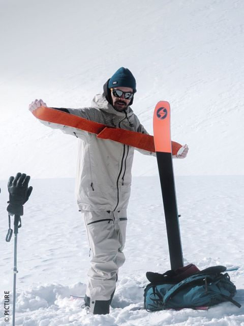Une personne en équipement d'hiver tire une peau de phoque orange sur un ski sur une montagne enneigée, tout en portant des bâtons de ski et un sac à dos.