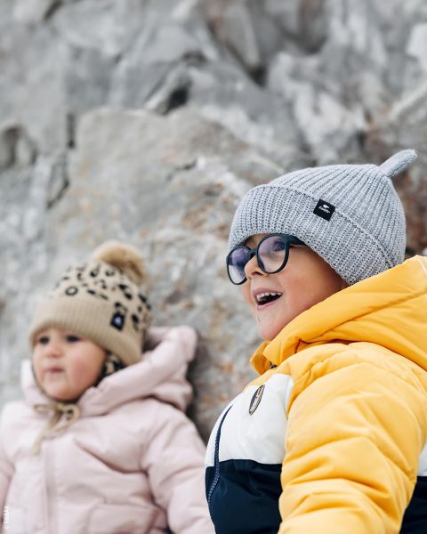Deux jeunes enfants en vestes d'hiver et bonnets en tricot sourient à l'extérieur, assis devant un fond rocheux. L'un porte du jaune, l'autre du rose.