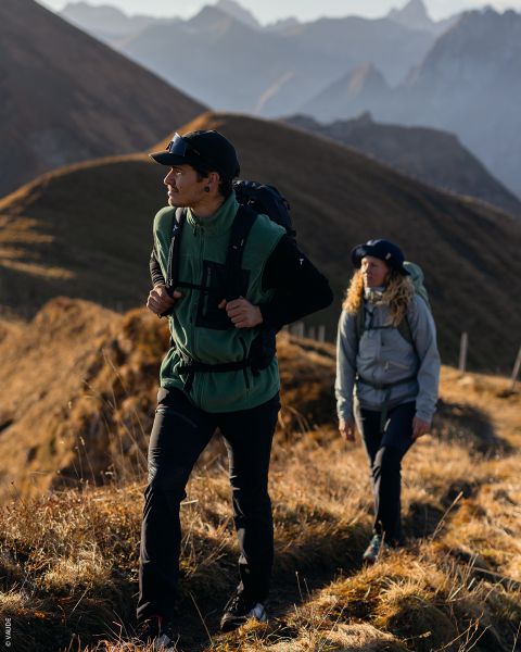 Deux randonneurs en équipement de plein air marchent sur un sentier de montagne ensoleillé avec des sommets escarpés et des collines herbeuses en arrière-plan.