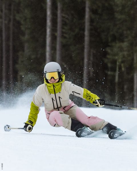 Un skieur avec un casque blanc, une veste jaune et un pantalon rose prend un virage serré sur une piste enneigée, projetant de la neige en l'air.