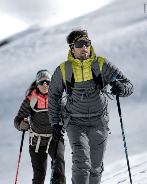 Deux personnes en équipement d'hiver montent une pente de montagne enneigée avec des bâtons de ski, des lunettes de soleil et des sacs à dos sous un ciel clair et nuageux.