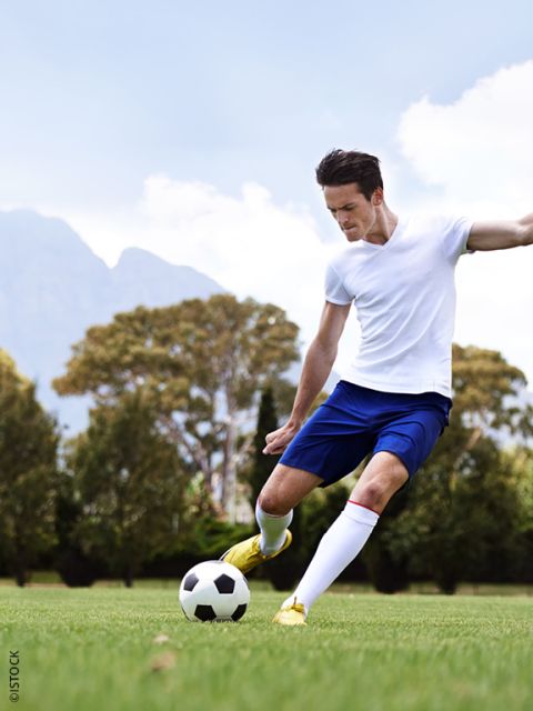 Un joueur de football en chemise blanche et short bleu frappe un ballon de football dans une prairie avec des arbres et des montagnes en arrière-plan.
