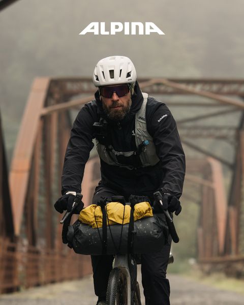 Un cycliste avec un casque et une chemise blanche se tient là avec son vélo et regarde au loin au coucher du soleil. À l'arrière-plan, on peut voir des arbres, des collines et une ligne électrique. Au-dessus, on peut lire le mot « ALPINA ».