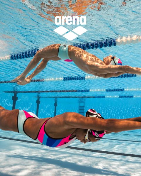 Un nageur portant un bonnet de bain noir et des lunettes de natation réfléchissantes glisse sous l'eau dans une piscine marquée par des séparateurs de couloir. Son maillot de bain bleu est orné de détails orange. L'eau est claire et le logo Arena est visible au-dessus.