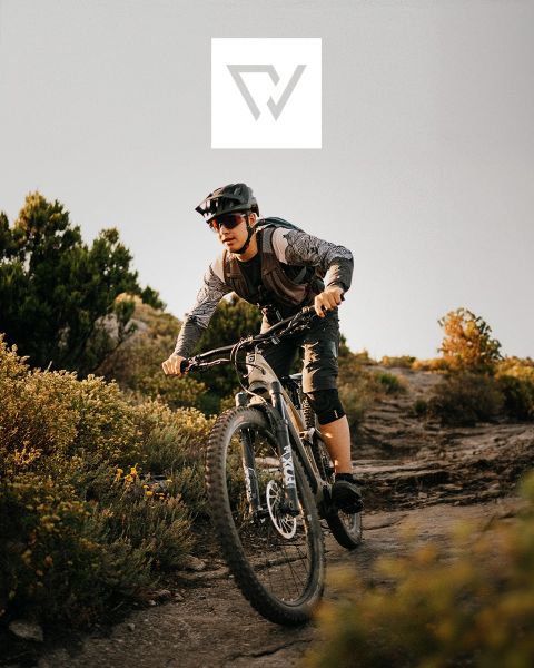 Deux cyclistes font du VTT sur un sentier accidenté dans une région montagneuse avec des sommets enneigés en arrière-plan. Ils portent des casques et des équipements de plein air. Un logo est visible en haut de l'image.
