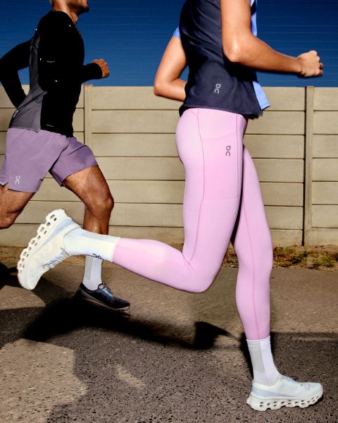 Une personne court à l'extérieur. Elle porte un coupe-vent beige-gris, un short gris clair et des chaussures de course blanches. L'arrière-plan est un ciel clair et une surface plane. Le logo « On » est visible dans le coin supérieur droit.