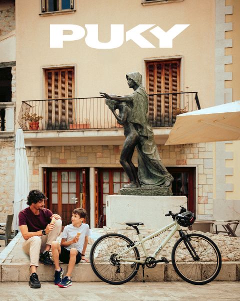 Une jeune fille avec un casque bleu et un t-shirt rayé fait du vélo bleu clair par une journée ensoleillée. Souriante, elle passe devant des graffitis colorés dans un environnement urbain. Le logo PUKY et le slogan « Ready for life » sont visibles en haut à gauche.