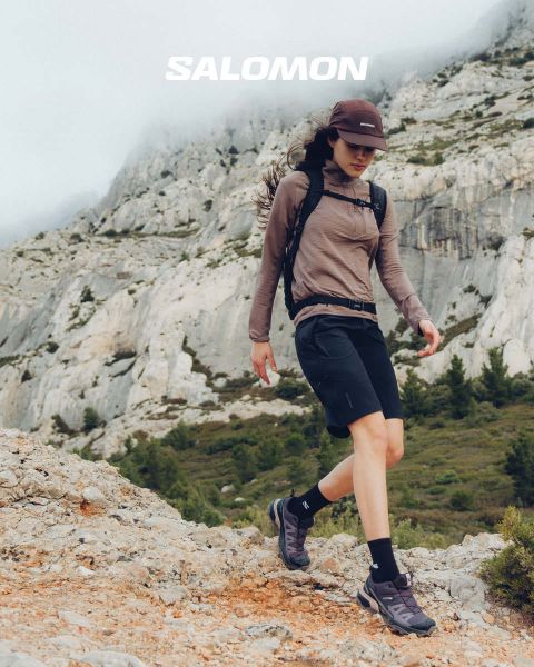Gros plan d'une personne portant des chaussures Salomon et des chaussettes Salomon avec un dégradé de couleurs bleues. Elle se tient dans une prairie avec un paysage de montagne pittoresque en arrière-plan. Un randonneur en vêtements d'extérieur se tient plus loin, également dans la prairie.