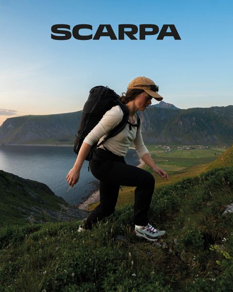 Une femme et un homme sont assis à l'extérieur sur de gros rochers. Ils portent des vêtements décontractés et des lunettes de soleil. Ils sont entourés de verdure et de rochers, le ciel est légèrement nuageux. En haut de l'image, on peut lire le mot « SCARPA ».
