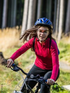 Une femme portant une chemise rouge à manches longues, un casque et des lunettes fait du vélo dans une zone herbeuse et boisée par une journée ensoleillée.