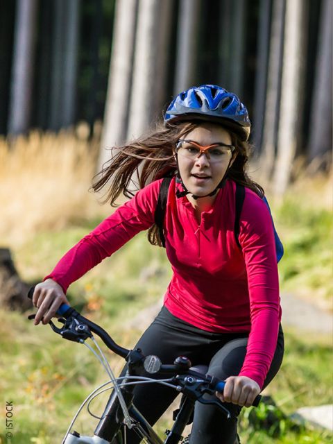Une femme portant une chemise rouge à manches longues, un casque et des lunettes fait du vélo dans une zone herbeuse et boisée par une journée ensoleillée.