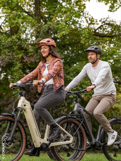 Une femme et un homme font du vélo électrique ensemble dans un parc verdoyant, tous deux portant des casques et des vêtements décontractés, souriant et profitant de la nature.