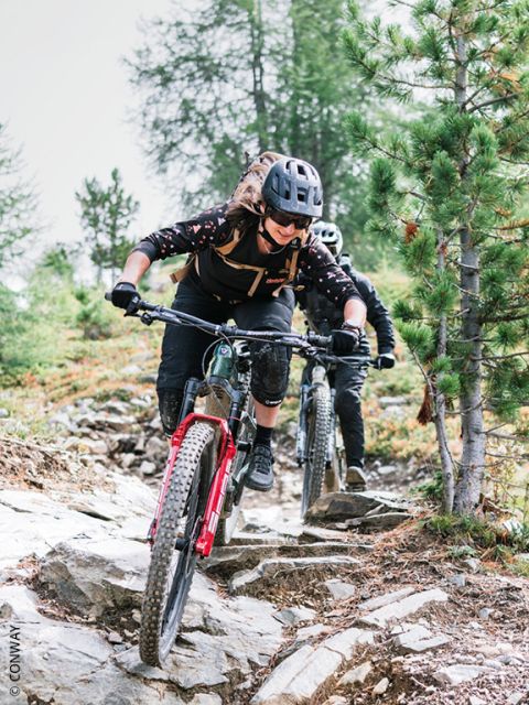 Deux cyclistes portant des casques descendent un chemin rocailleux et boisé avec leurs VTT, se concentrant et naviguant entre les arbres et le terrain accidenté.