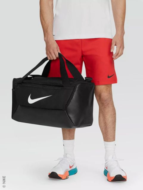 Une personne portant un short Nike rouge, une chemise blanche et des baskets blanches tient un sac de sport Nike noir avec un logo Swoosh blanc.
