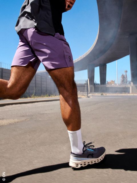 Personne marchant dans une rue ensoleillée sous un viaduc d'autoroute, portant un short violet, des chaussettes blanches et des chaussures de course grises.