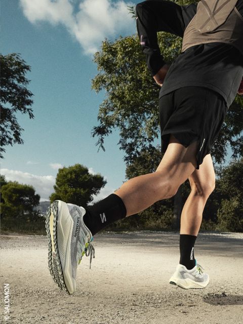 Un coureur en short noir et veste et baskets blanches Salomon fait du jogging sur un chemin de gravier, avec des arbres et un ciel bleu en arrière-plan.