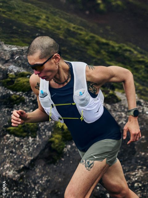 Une personne tatouée marche sur un chemin rocailleux et mousseux en montée, portant des lunettes de soleil, un gilet d'hydratation et une montre connectée.