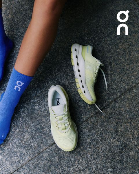 Une personne en chaussettes bleu clair à côté d'une paire de chaussures de course ON jaune clair sur un sol en pierre grise, avec le logo ON dans le coin supérieur droit.