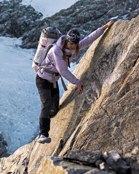 Une femme avec un sac à dos escalade une pente de montagne raide et rocheuse. Elle porte un équipement de plein air et un chapeau, tandis que des sommets enneigés sont visibles en arrière-plan.