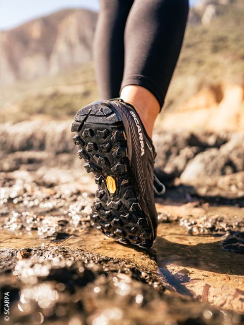 Gros plan d'une personne portant des chaussures de trail noires courant sur un sol rocheux et humide à l'extérieur, avec des montagnes en arrière-plan flou.