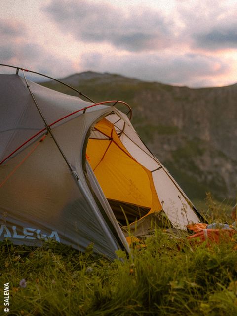 Une tente grise et jaune installée dans une prairie au coucher du soleil, avec des montagnes en arrière-plan.