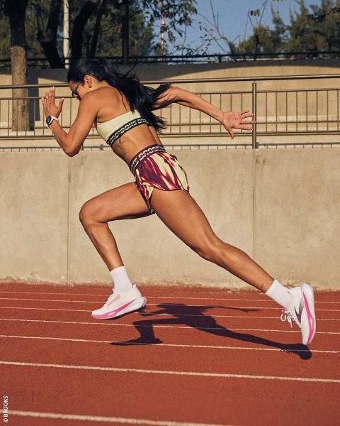 Une femme en tenue de sport sprinte sur une piste de course rouge en plein air, les bras qui pompent et le corps penché vers l'avant au milieu de la foulée.