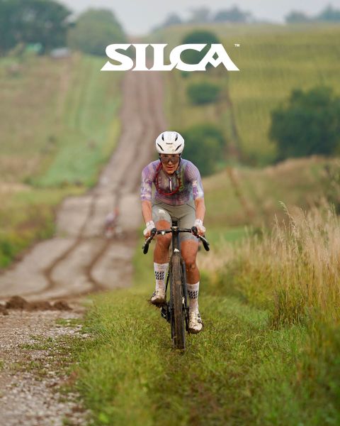 Un cycliste monte une route de gravier à travers des champs herbeux. Il porte un casque et une tenue de cycliste, avec le logo "SILCA" en haut de l'image.