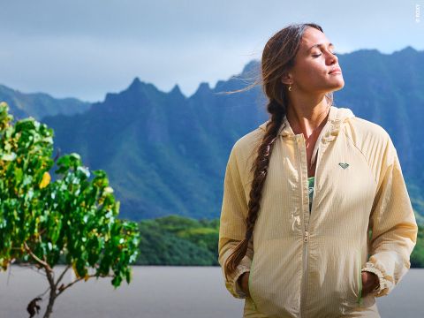 Une femme vêtue d'une veste beige se tient au bord d'un lac avec des montagnes en arrière-plan, les yeux fermés et le visage tourné vers le soleil, à côté d'un arbre feuillu.