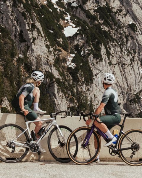 Deux cyclistes en tenue assortie s'arrêtent à une barrière de route de montagne, avec des falaises rocheuses et des plaques de neige en arrière-plan.