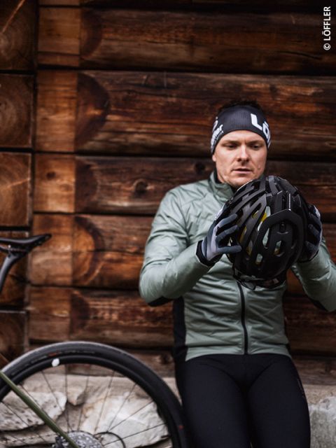 Un cycliste en veste verte et bonnet noir inspecte un casque tout en s'appuyant contre un mur en bois à côté d'un vélo.