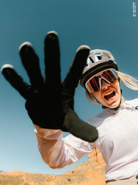 Un cycliste avec un casque et des lunettes de soleil sourit énergiquement et tend une main gantée vers la caméra devant un ciel bleu clair.