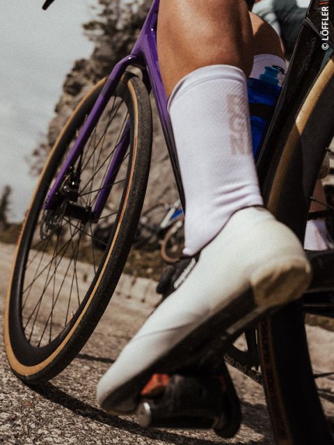Gros plan sur la jambe et la chaussure blanche d'un cycliste sur un vélo violet, avec une route floue et un paysage rocheux en arrière-plan.