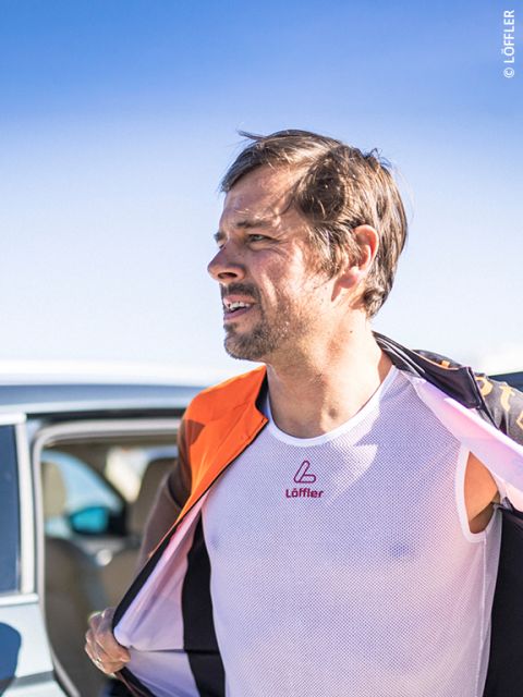 Un homme à l'extérieur, portant un maillot de corps blanc et une veste orange, se tient à côté d'une voiture sous un ciel bleu clair.