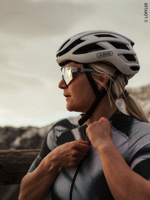 Une cycliste portant un casque et des lunettes de soleil ajuste son maillot à l'extérieur, avec des montagnes en arrière-plan au coucher du soleil.