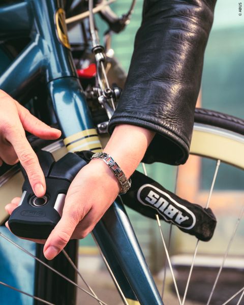 Une personne en veste de cuir attache un vélo bleu avec un antivol de vélo ABUS, en se concentrant sur la sécurisation du cadre et de la roue.