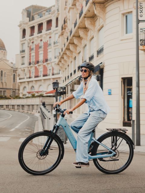 Une femme avec un casque et des lunettes de soleil roule sur un vélo bleu dans une rue bordée d'élégants bâtiments.