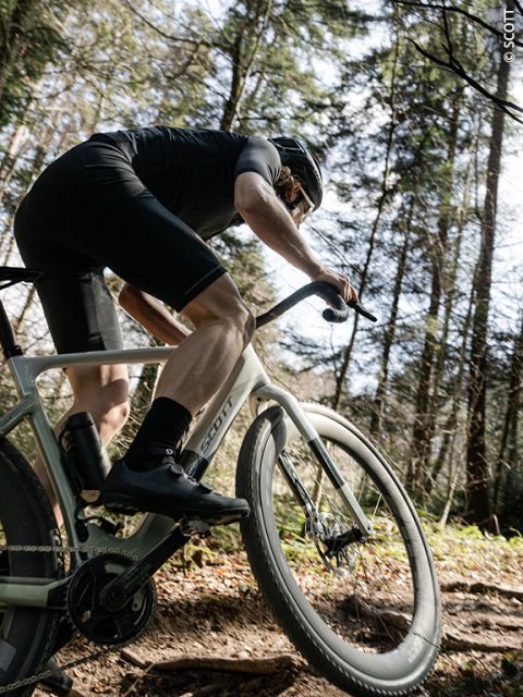 Un cycliste vêtu de noir monte une côte sur un chemin de terre avec un vélo de gravel, à travers une forêt ensoleillée.