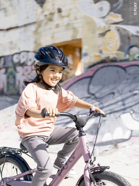 Une jeune fille avec un casque sourit en faisant du vélo rose devant un graffiti coloré par une journée ensoleillée.