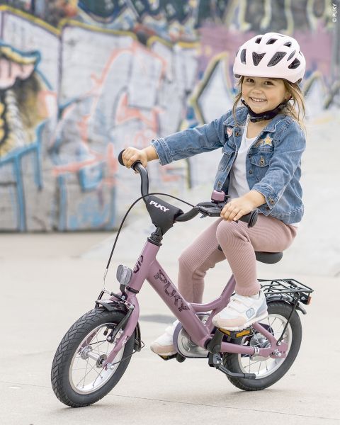 Une jeune fille avec un casque rose et une veste en jean sourit en faisant du vélo violet devant un mur couvert de graffitis.