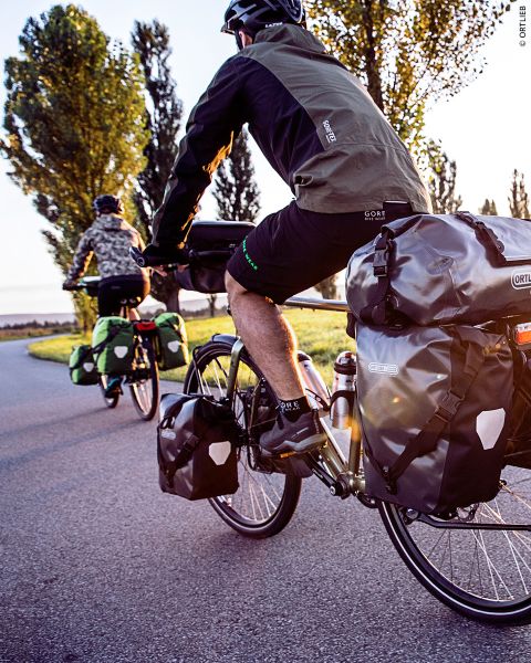 Deux cyclistes avec des sacoches roulent sur une route pavée entourée d'arbres et baignée d'une chaude lumière du soleil.