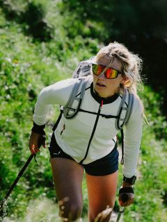 Une femme marche en montée sur un sentier verdoyant, portant des lunettes de soleil, une veste blanche, un sac à dos et utilisant des bâtons de trekking.