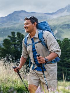 Un homme souriant aux cheveux longs et à la barbe, vêtu d'une chemise bleue et d'un sac à dos, se tient à l'extérieur devant un arrière-plan naturel flou.